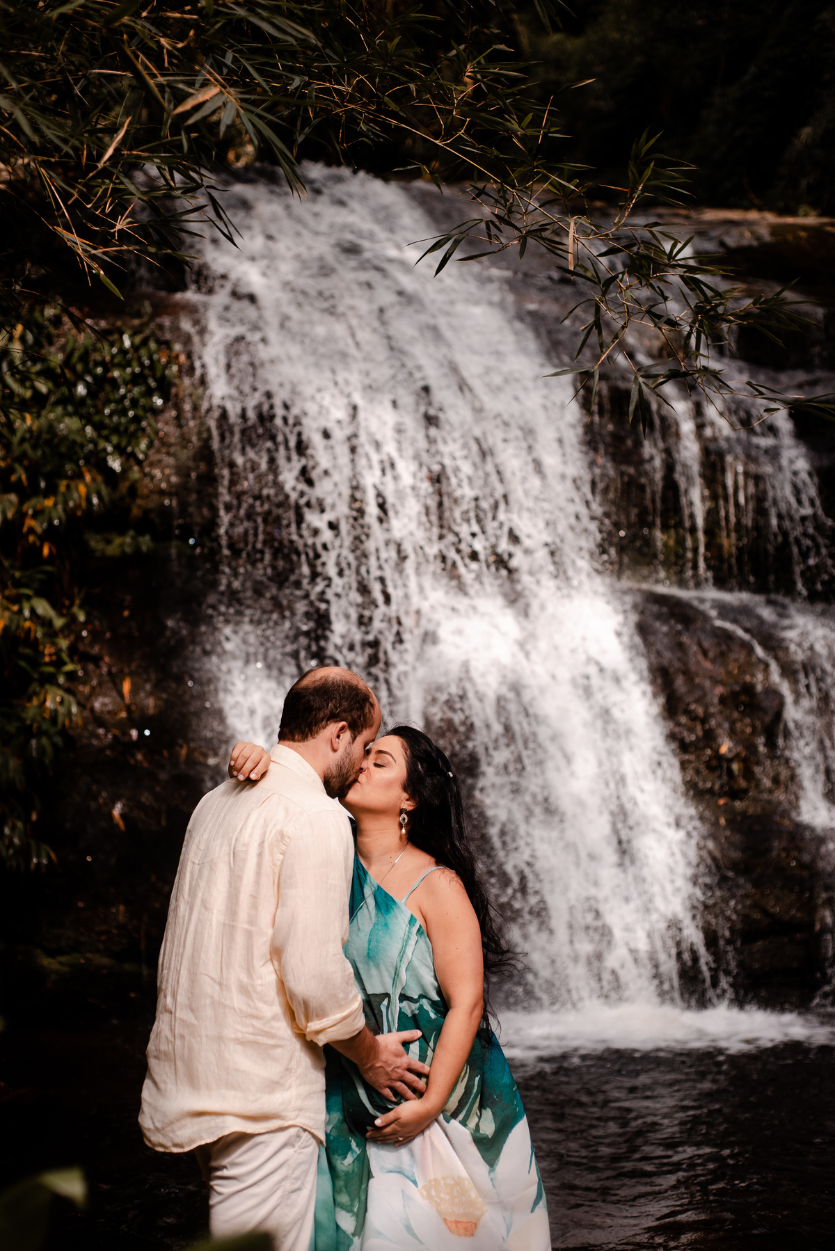 ensaio externo de gestante na cachoeira em nova friburgo, região serrana do rio de janeiro. Foto da gestante e o companheiro dando um beijo na frente da queda d'água