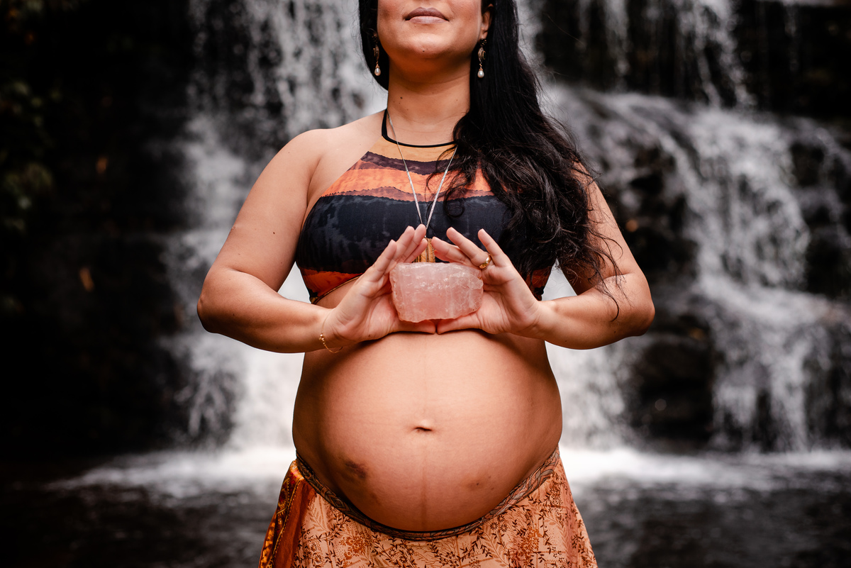 ensaio externo de gestante na cachoeira em nova friburgo, região serrana do rio de janeiro. Foto da gestante em frente da queda d'água da cachoeira segurando um cristal rosa com as mãos