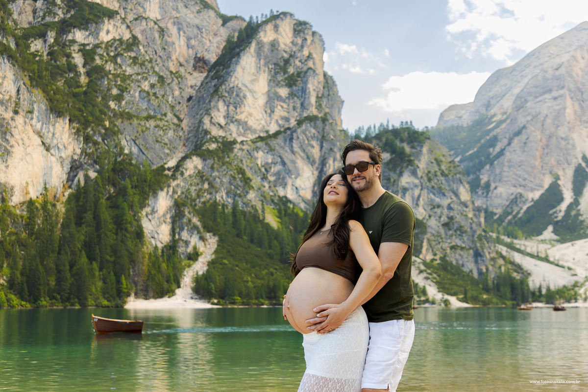 Ensaio fotográfico de gestante com fotógrafo de Milão brasileiro e casal abraçado no Lago di Braies com montanhas das Dolomitas ao fundo e lago azul e verde com barcos na água