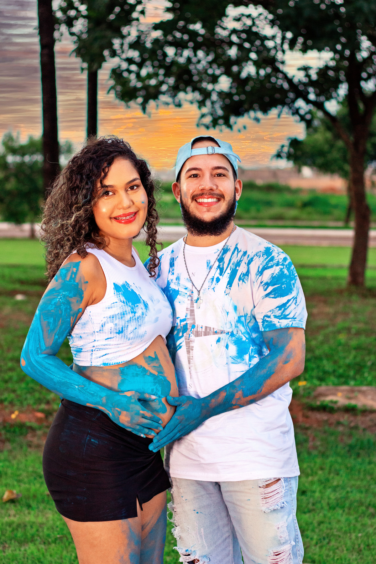 Jainne e seu parceiro em um cenário romântico, com roupas brancas manchadas de tinta azul. O pôr do sol reflete a emoção e a expectativa da chegada do bebê.