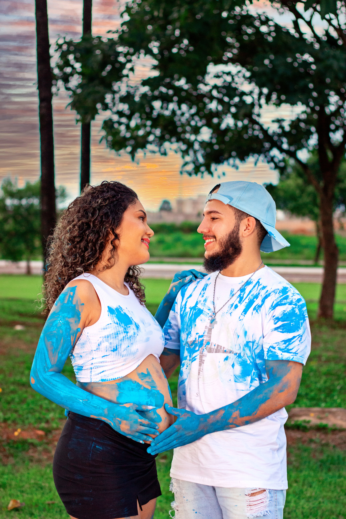 Jainne e seu parceiro vestindo roupas brancas, sorrindo enquanto se cobrem de tinta azul em uma celebração vibrante da revelação do sexo do bebê. O pôr do sol ao fundo cria um cenário mágico.