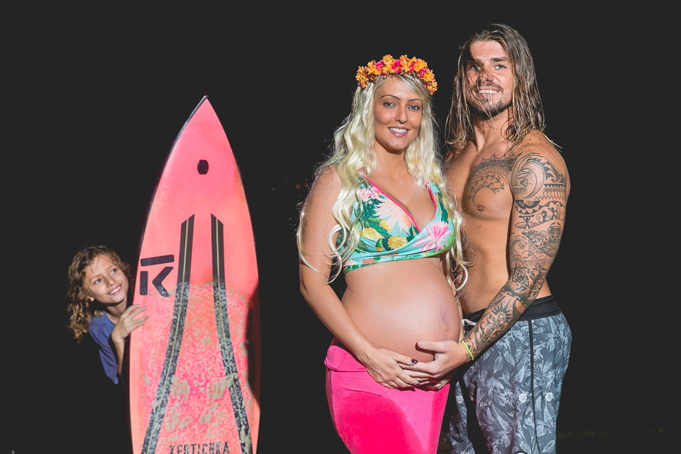 Casal que curte um surfe, e o irmão mais velho espiando atrás da prancha. 
