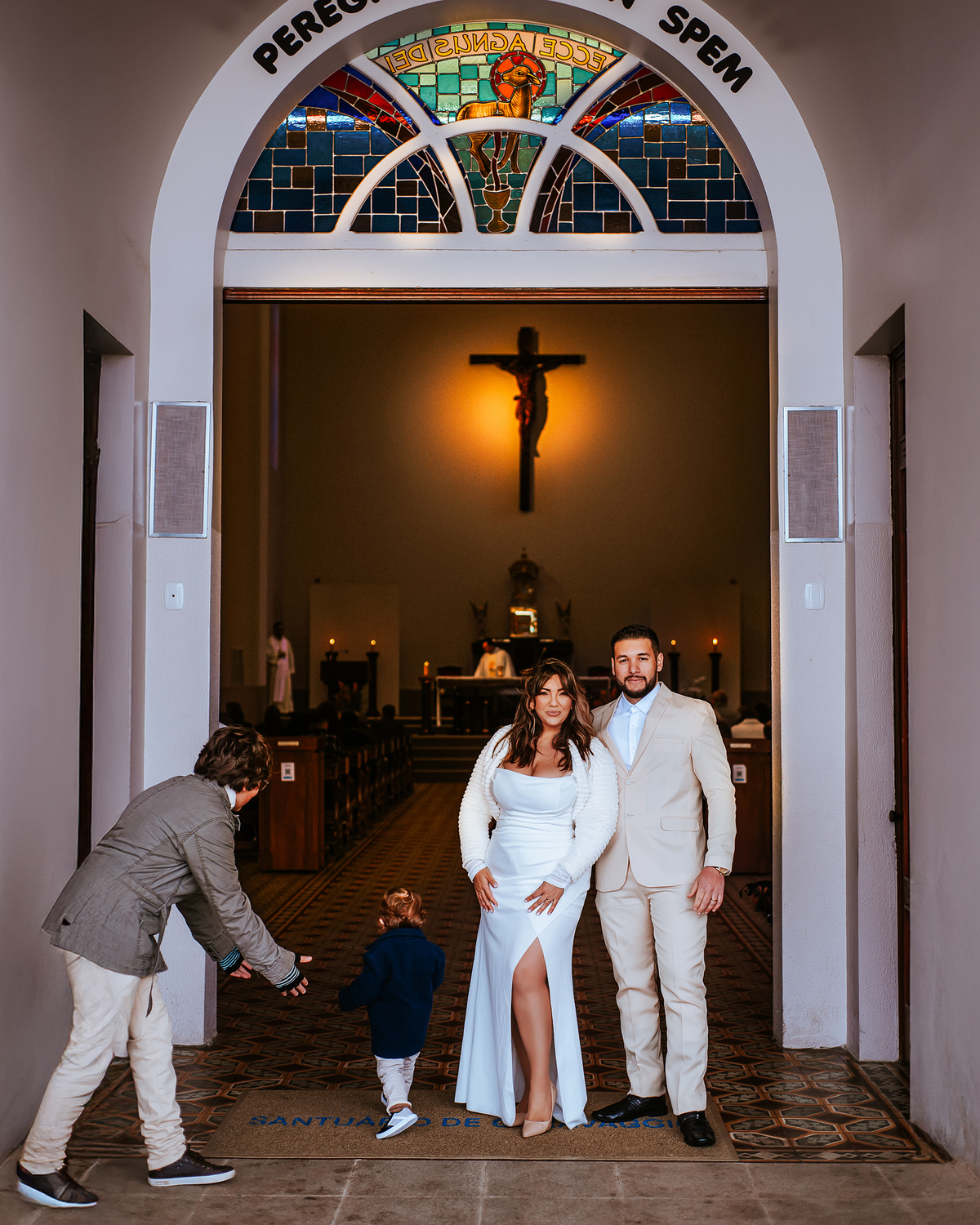 Casal recém-casado fotografado na entrada de uma igreja, com o altar e o crucifixo ao fundo.