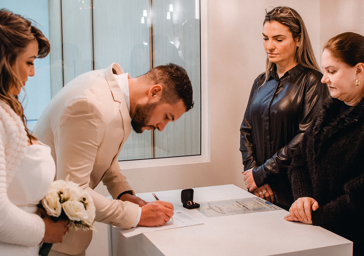 Noivo assina documentos durante cerimônia de casamento civil no cartório de Farroupilha, acompanhado pela noiva e testemunhas.