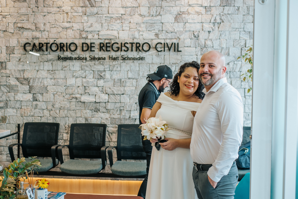 Stefhanie e John abraçados em retrato no local da cerimônia civil, com parede de pedra ao fundo.