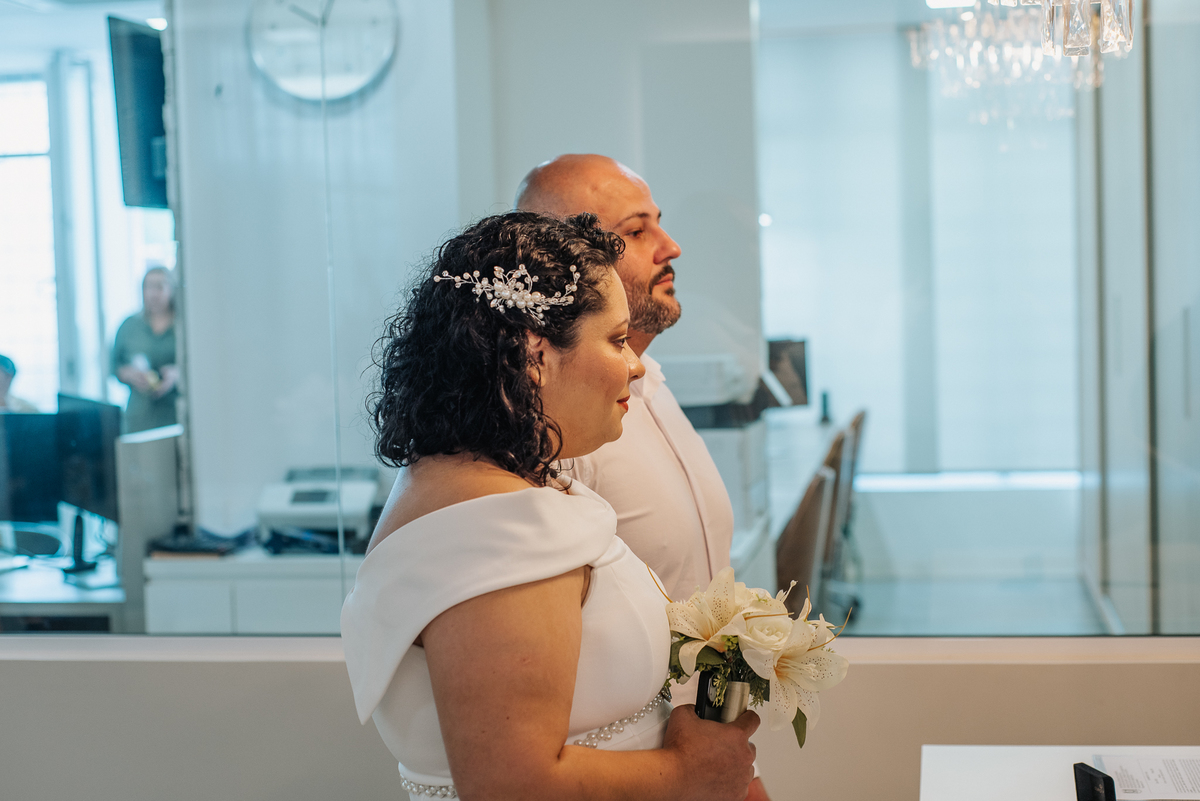Stefhanie aparece de perfil segurando seu buquê de flores brancas durante a cerimônia de casamento civil.