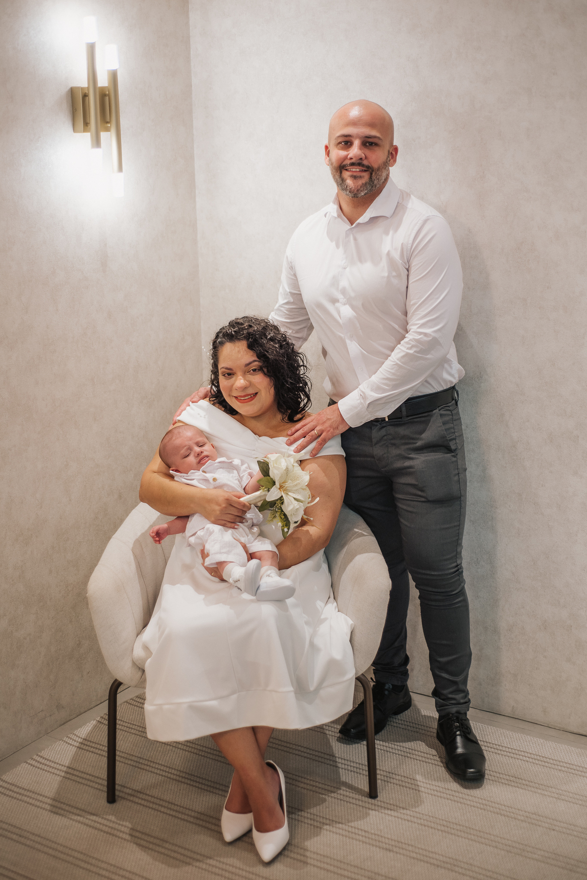 Stefhanie sentada com o bebê no colo e o buquê de flores brancas, acompanhada por John.