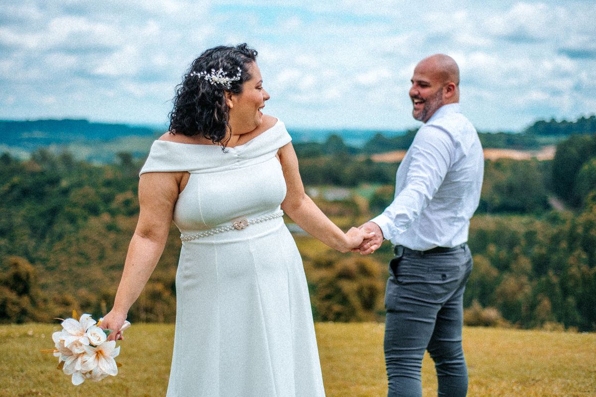Stefhanie segura o buquê enquanto está de mãos dadas com John em uma paisagem verde da Serra Gaúcha.