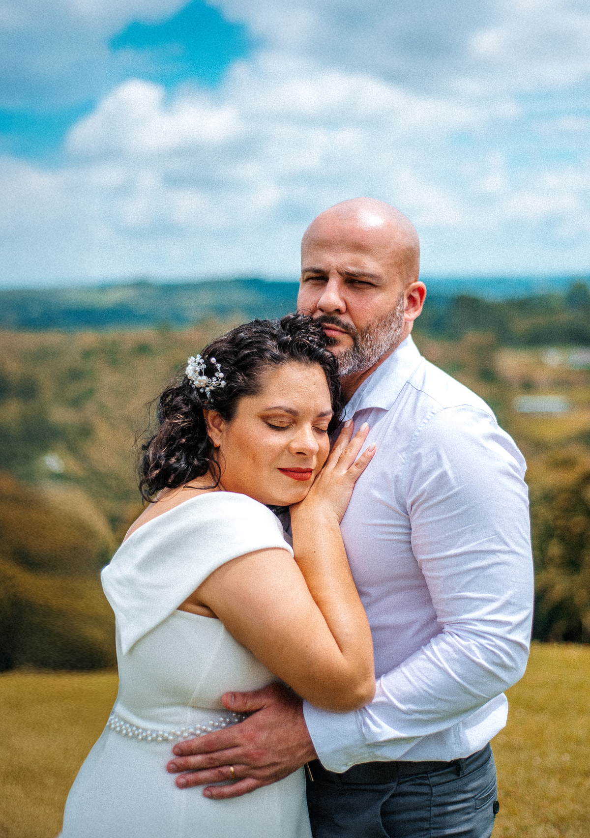 Stefhanie abraça John com os olhos fechados durante ensaio externo de casamento com paisagem verde ao fundo.