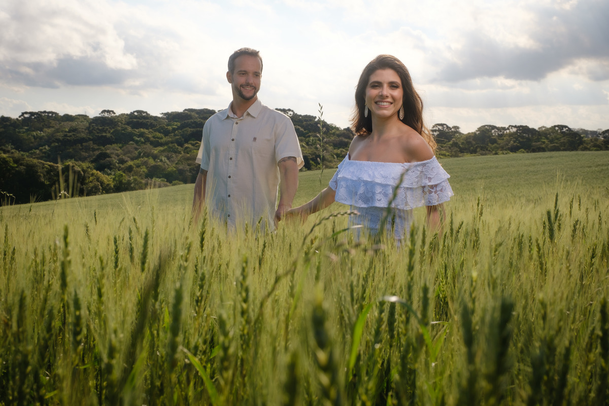 Ensaio de casal no Recanto Nogarotto em São José dos Pinhais com o noiva em primeiro plano de mão dada ao noivo em segundo plano em fotografia ao ar livre em meio ao campo de trigo