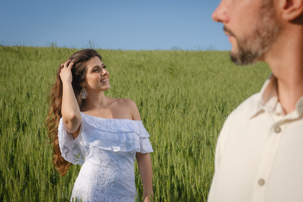 Fotografia do ensaio de casal em campo de trigo ao ar livre aparecendo em primeiro plano o noivo em corte ao lado direto da foto emoldurando a noiva que aparece em segundo plano mexendo nos cabelos no Recanto Nogarotto em São José dos Pinhas