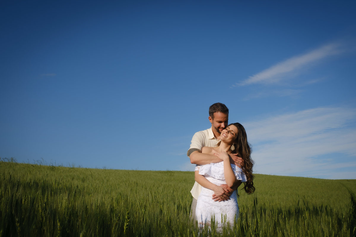 Noivo abraça a noiva por trás em fotografia ao ar livre em meio ao campo de trigo no Recanto Nogarotto em São José dos Pinhais onde eles sorriem de olhos fechados curtindo o momento