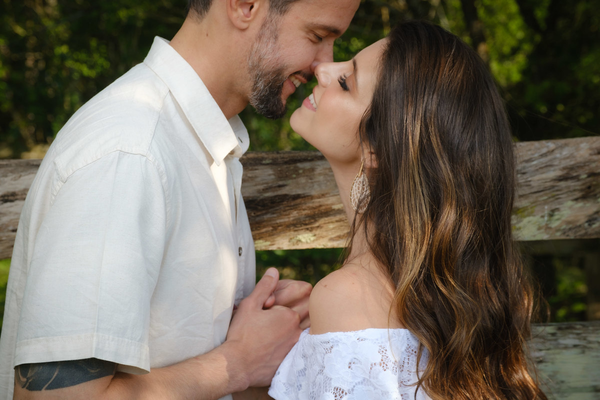 Noiva de frente e segurando as mãos do noivo com os lábios próximos de se se beijarem e sorrindo um para o outro em ensaio de casal no Recanto Nogarotto em São José dos Pinhais