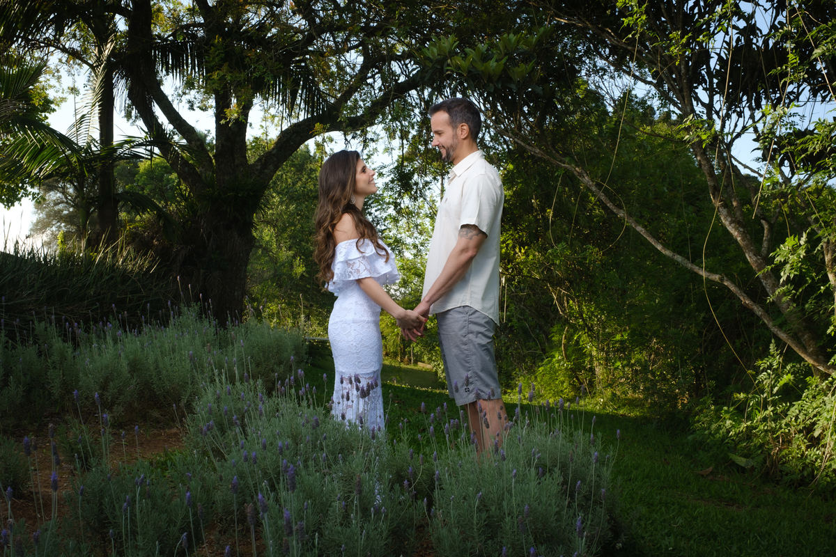 Plantação de lavandas em primeiro plano com os noivos logo em segundo plano de frente um para o outro e de mãos dada com as árvores que emolduram os noivos em terceiro plano no Recanto Nogarotto em São José dos Pinhais