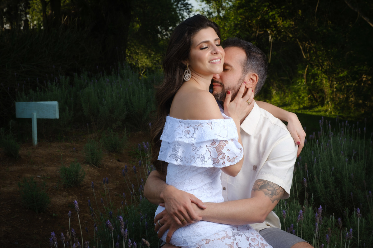 Ensaio de casal no Recanto Nogarotto em São José dos Pinhais com o noivo sentado abraçando a noiva pela cintura que está sentada na perna direta do noivo com o braço esquerdo sobre o ombro do noivo e a mão direita acariciando a barba do noivo