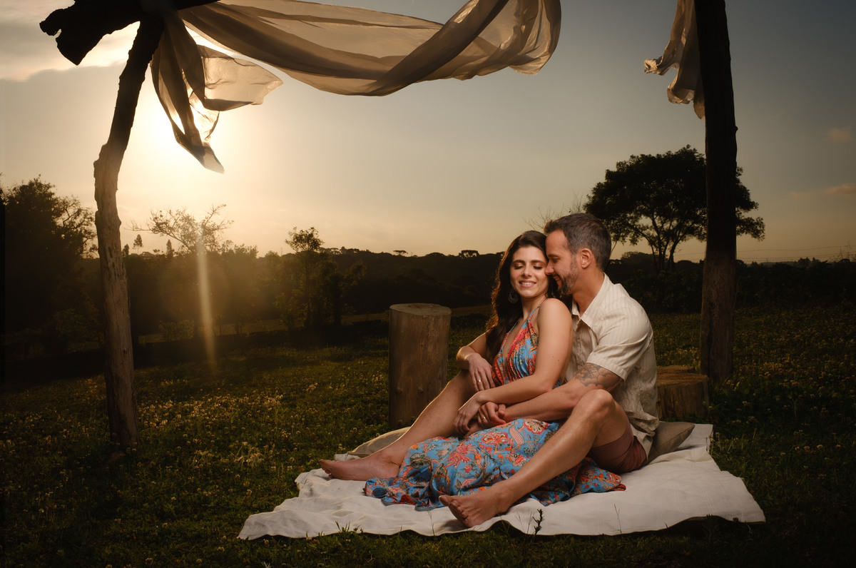 Noivos sentados sobre uma manta no chão e sob um gazebo feito com troncos de madeira e com voal branco com o por do sol ao fundo no Recanto Nogarotto em São José dos PInhais