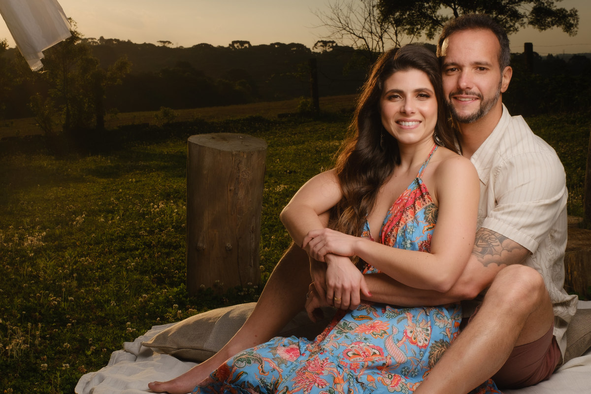 Noivos olhando para a fotos com a noiva sentada no chão  e entre as pernas do noivo que a abraça pela cintura em ensaio de casal no Recanto Nogarotto em São José dos Pinhais