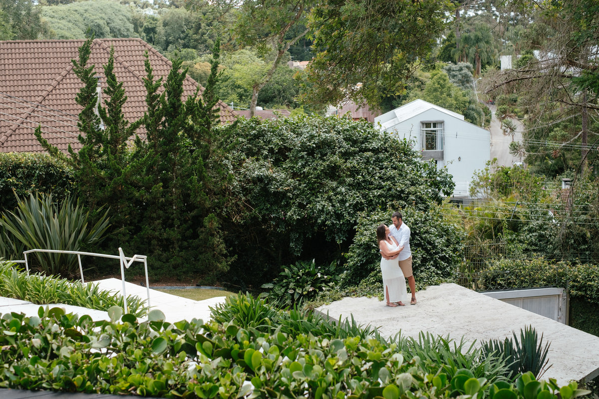 Vista ampla com os noivos abraçados aparecendo no terço inferior sobre uma base de concreto com plantas verdes no primeiro plano da foto e a área verde do ambiente ao fundo da imagem