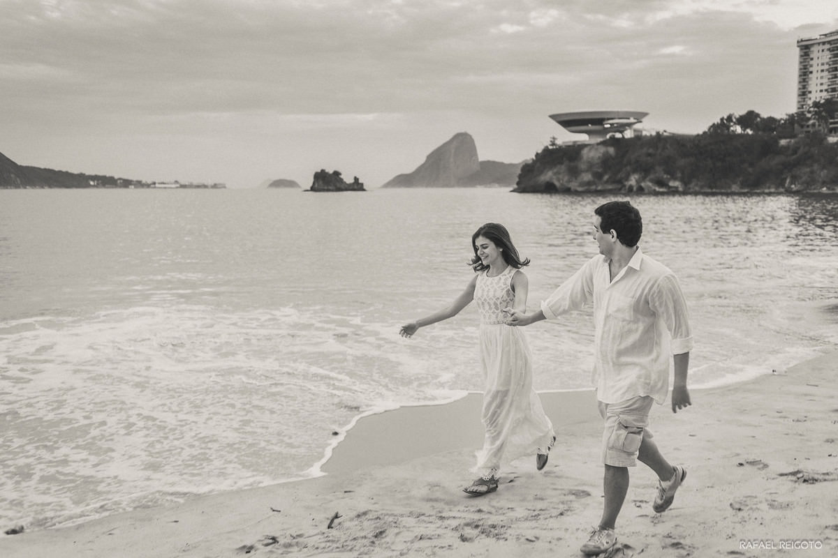 O casal Mariana e Israel caminhando descontraído no ensaio pré-casamento, Praia de Icaraí, Niterói/RJ