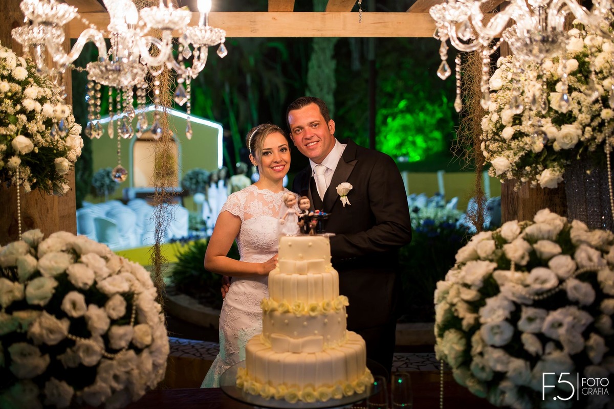 Noivos Lívia e Renato, sorrindo junto ao bolo em sua festa de casamento, em Muzambinho - MG.