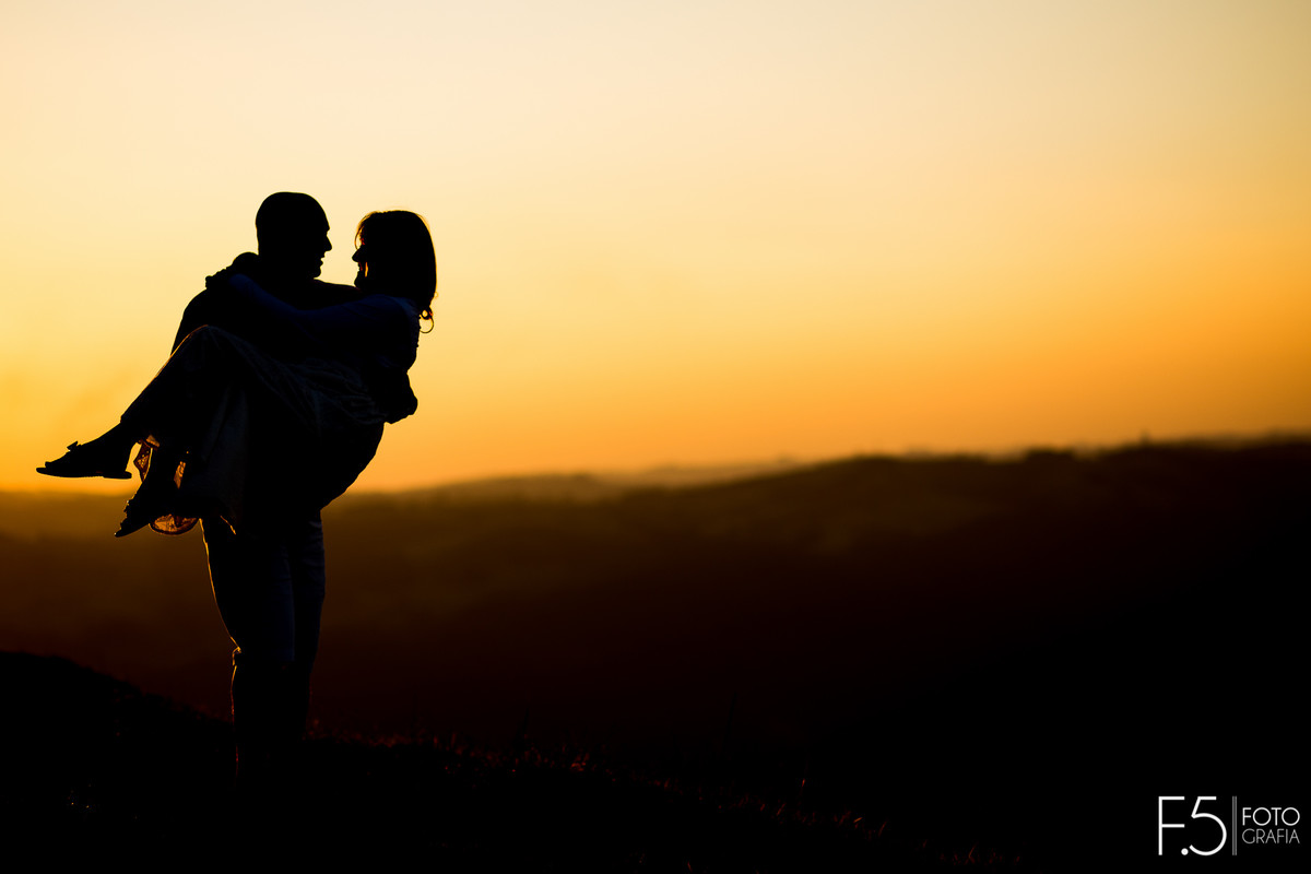 Homem segurando gestante no colo em meio ao pôr do sol, Ensaio Gestante Poços de Caldas - MG