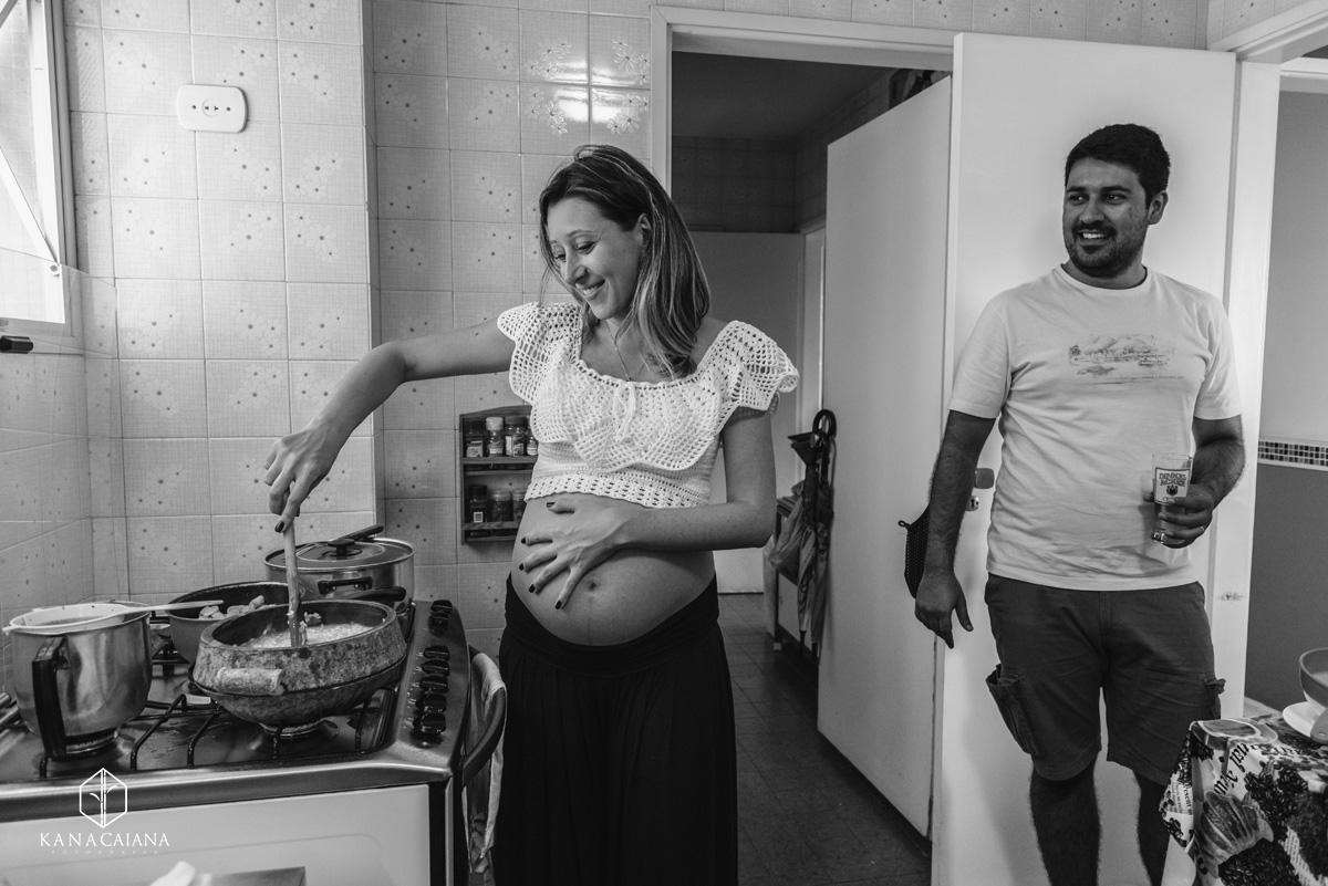 mulher grávida cozinhando e fazendo carinho na barriga enquanto marido sorridente observa durante ensaio de fotos de gestante em São Paulo sp