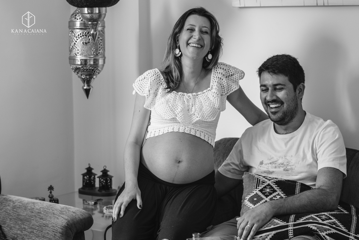 Mulher grávida e seu marido caem na gargalhada no sofá de casa durante ensaio de fotos de gestante em São Paulo sp