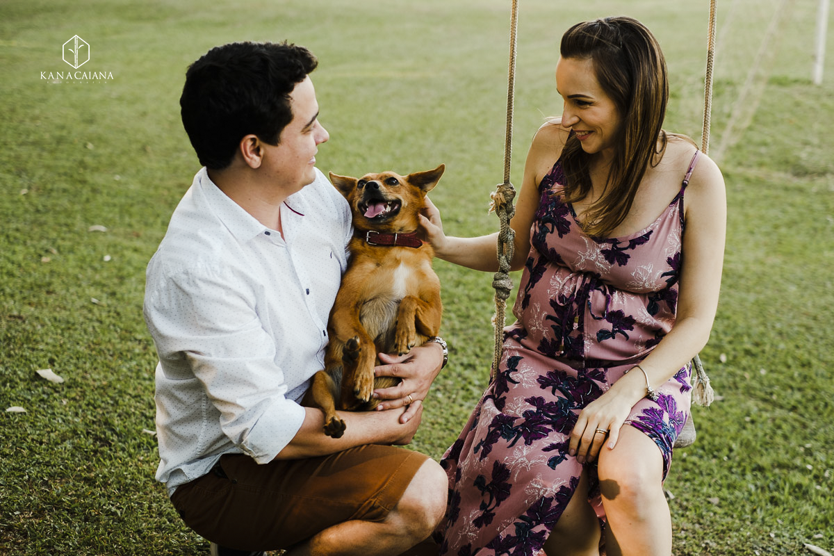 cachorro sorrindo para seu dono enquanto esposa gestante posa para ensaio de fotos no campo