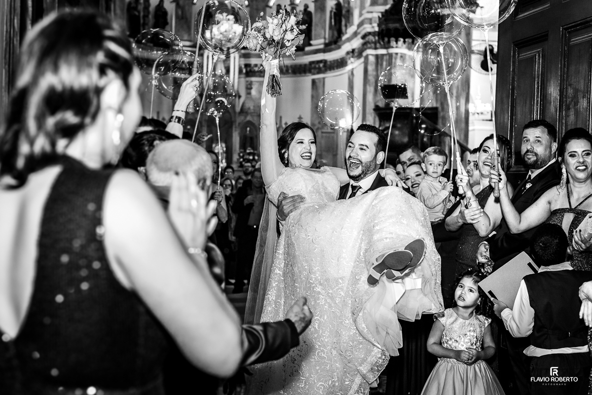  Noivo levando a noiva no colo durante a saída da Igreja, celebrando seu Casamento realizado no Santuário de São Benedito em Lorena/SP 