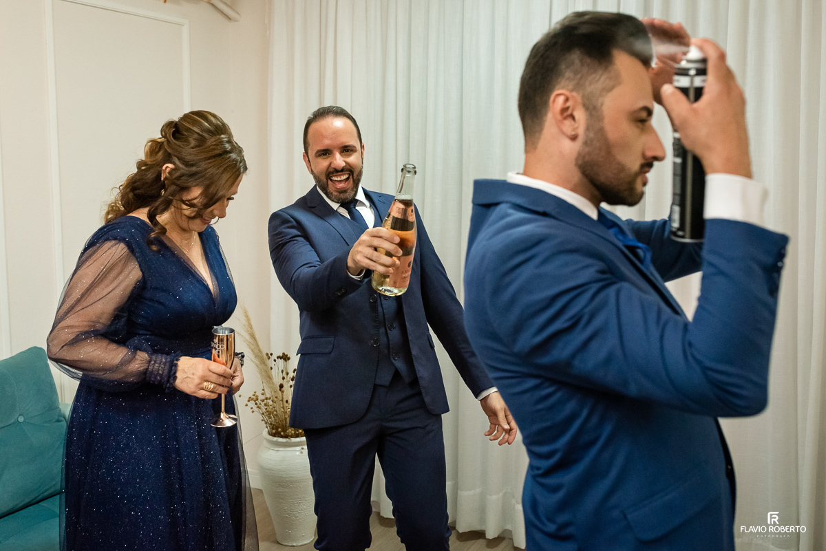 Noivo se preparando para abrir a Champagne com sua mãe, enquanto o outro noivo arruma o cabelo durante o making of no Espaço Luxo em Taubaté 