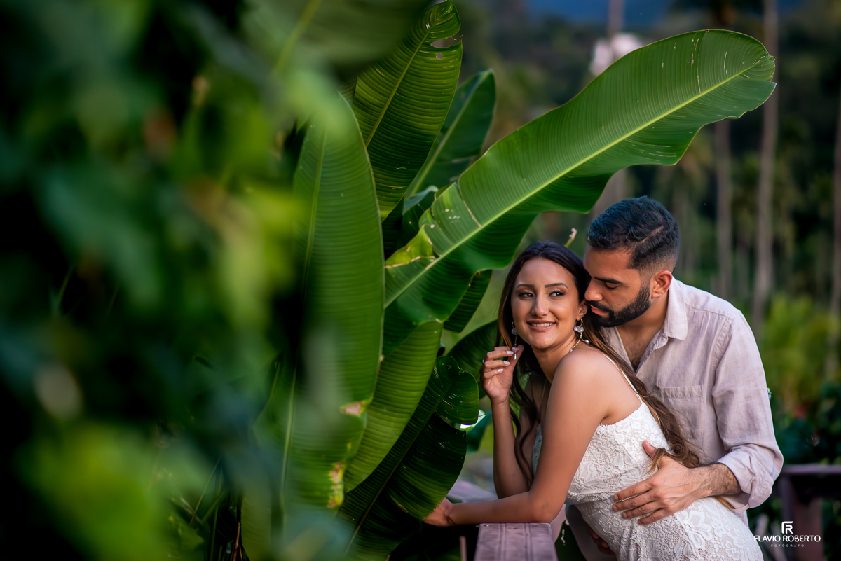 noivo abraçando sua noiva em um deck cheio de folhas de bananeira durante ensaio pre wedding em Ilhabela