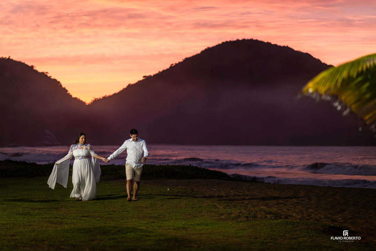 Casal Nathane e Igor correndo durante ensaio pre wedding na Praia Vermelha do Norte em Ubatuba, em frente a um pôr do sol com céu colorido em tons de azul, rosa e laranja, abraçados ao lado de um coqueiro, fotografados por Flavio Roberto Fotógrafo