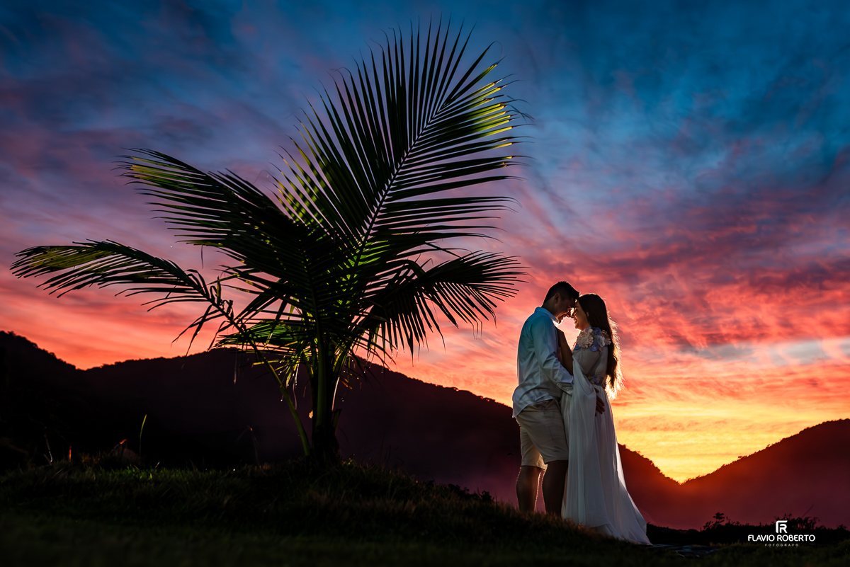 Casal Nathane e Igor durante ensaio pre wedding na Praia Vermelha do Norte em Ubatuba, em frente a um pôr do sol com céu colorido em tons de azul, rosa e laranja, abraçados ao lado de um coqueiro, fotografados por Flavio Roberto Fotógrafo
