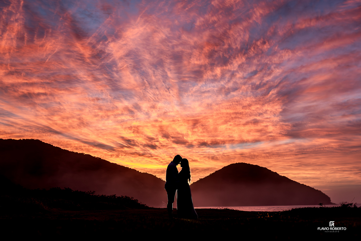 Casal Nathane e Igor durante ensaio pre wedding na Praia Vermelha do Norte em Ubatuba, em frente a um pôr do sol com céu colorido em tons de azul, rosa e laranja, abraçados ao lado de um coqueiro, fotografados por Flavio Roberto Fotógrafo