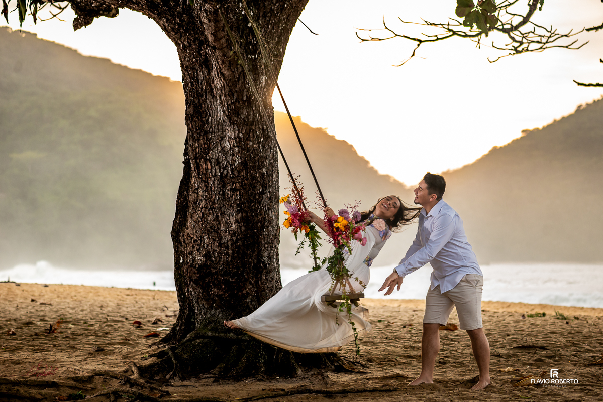 Momento de alegria e conexão entre o casal durante ensaio pré-wedding na Praia Vermelha do Norte, Ubatuba — a noiva no balanço florido, ao pôr do sol, sendo empurrada pelo noivo.