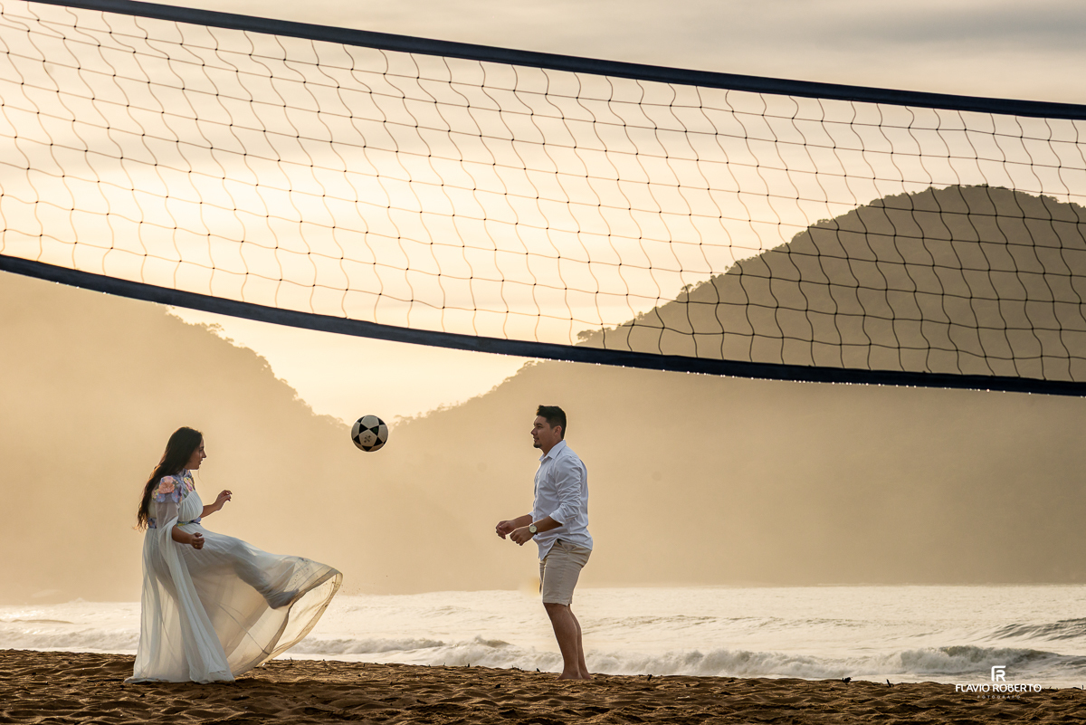 Casal brincando de altinha na rede de vôlei na Praia Vermelha do Norte, Ubatuba, com cenário de montanhas, mar e luz dourada do amanhecer.