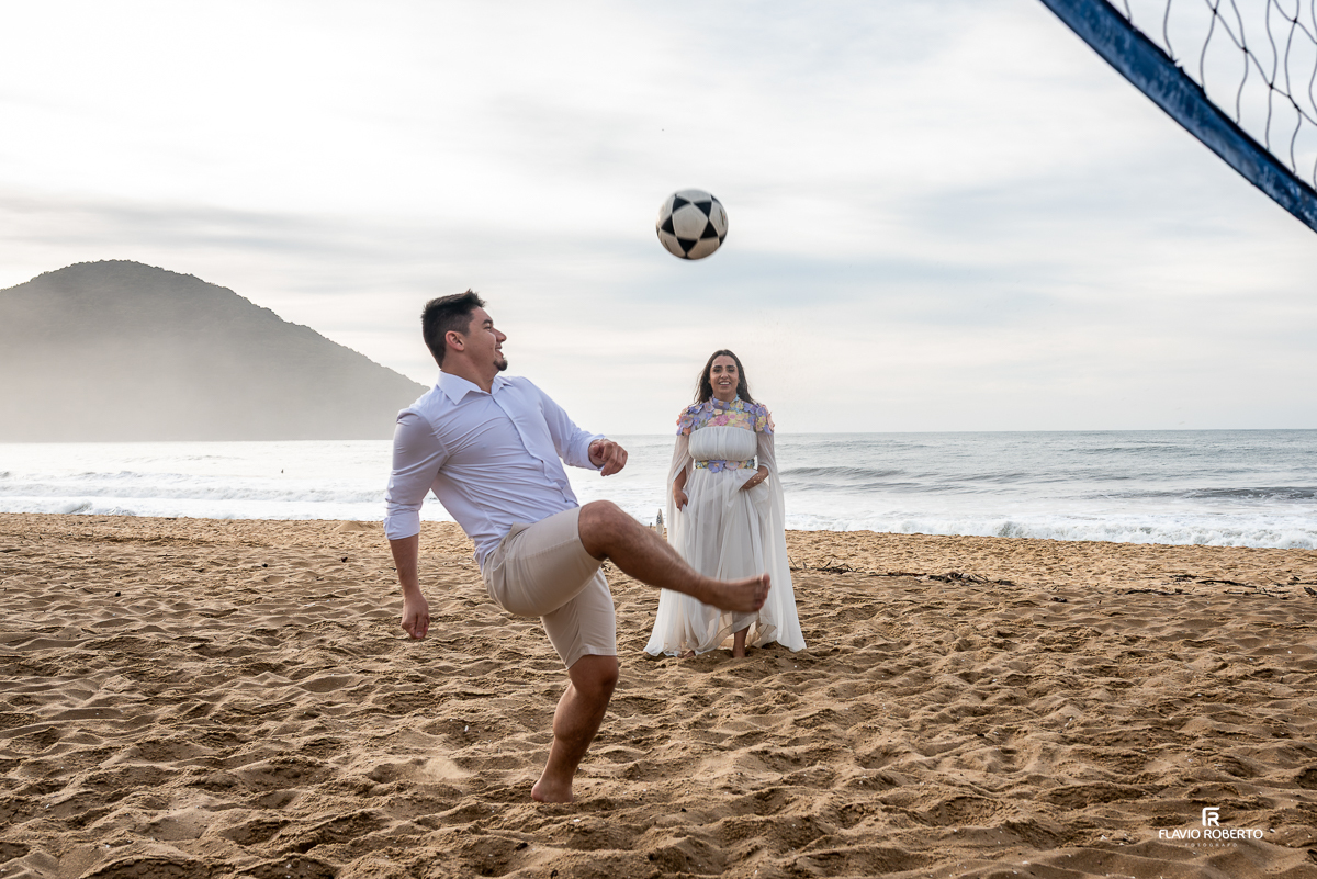 Casal brincando de altinha na rede de vôlei na Praia Vermelha do Norte, Ubatuba, com cenário de montanhas, mar e luz dourada do amanhecer.