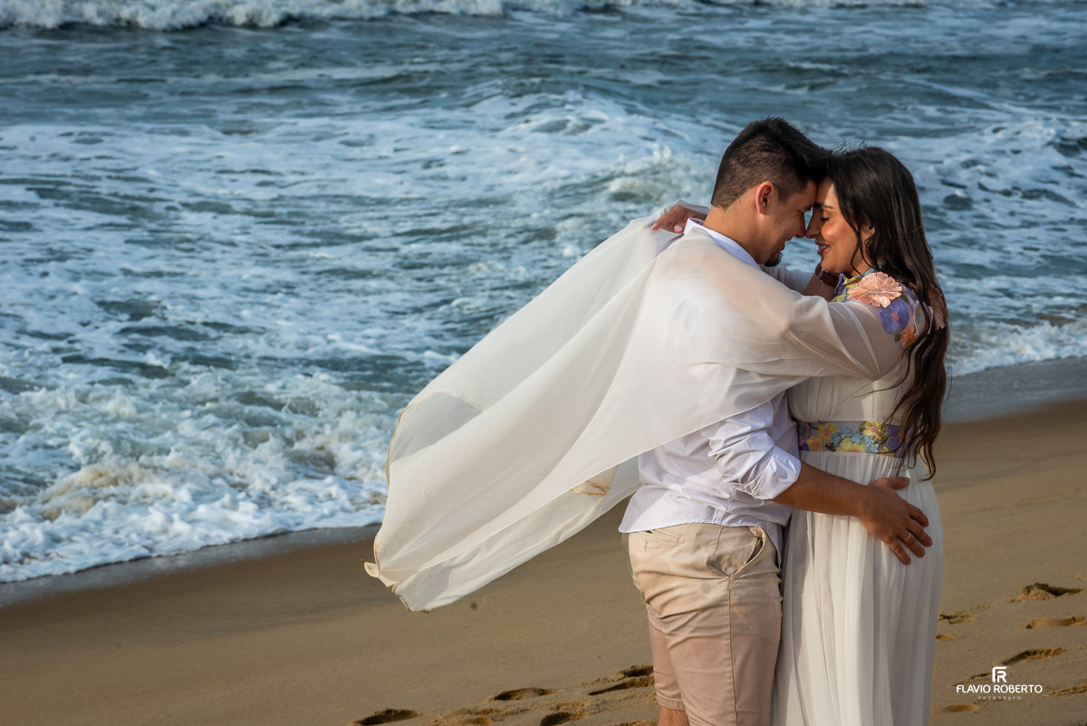 Casal se abraçando carinhosamente à beira-mar, com os pés na areia e ondas ao fundo, durante ensaio pré-wedding na Praia Vermelha do Norte, em Ubatuba.
