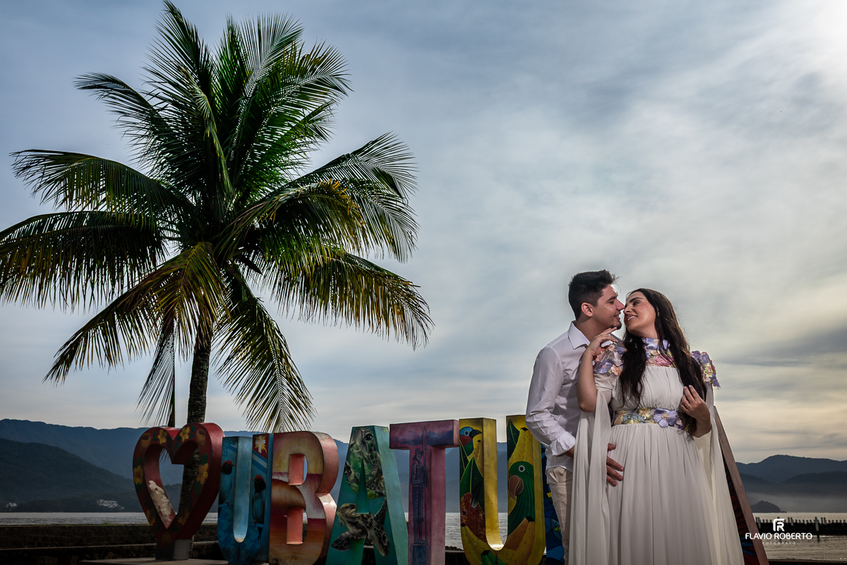 Casal apaixonado durante o ensaio pré-wedding na Praia Vermelha do Norte, em Ubatuba, ao pôr do sol, abraçados sob um coqueiro, com céu colorido em tons de azul, laranja e rosa.