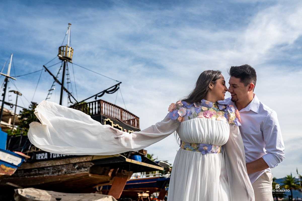 Casal apaixonado em ensaio pré-wedding em Ubatuba, diante de um barco pirata decorativo sob um céu azul, com a noiva de vestido esvoaçante com flores bordadas.