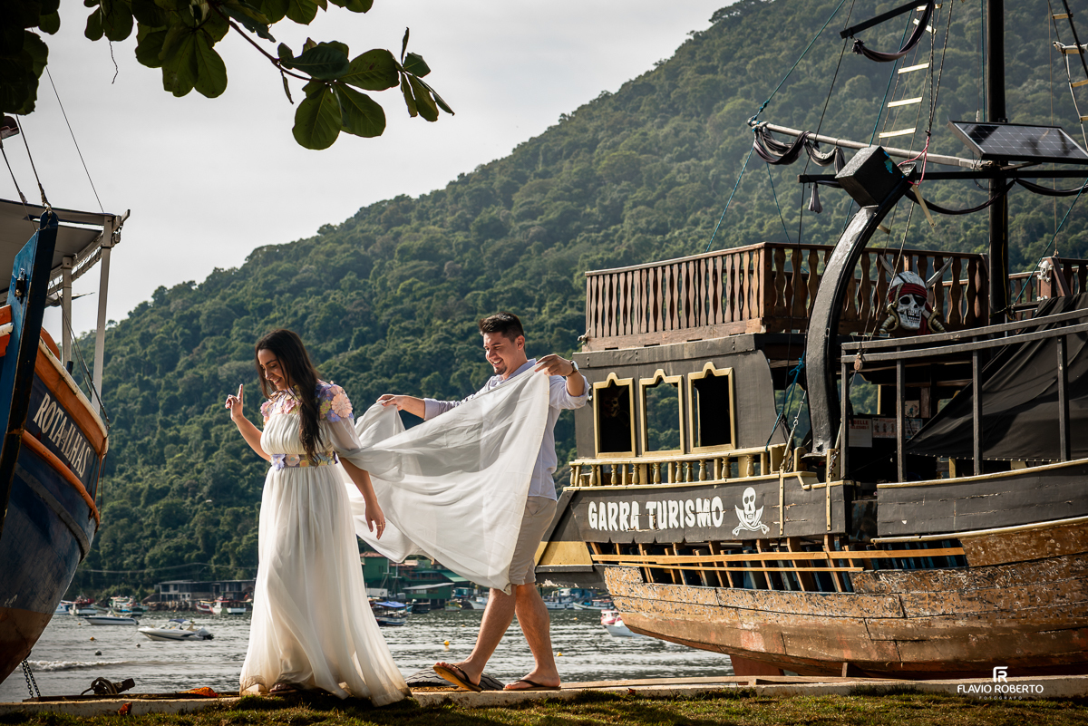 Casal caminha entre barcos em Ubatuba, com o noivo segurando com carinho o vestido da noiva, em um gesto espontâneo e romântico diante de um barco temático de pirata.