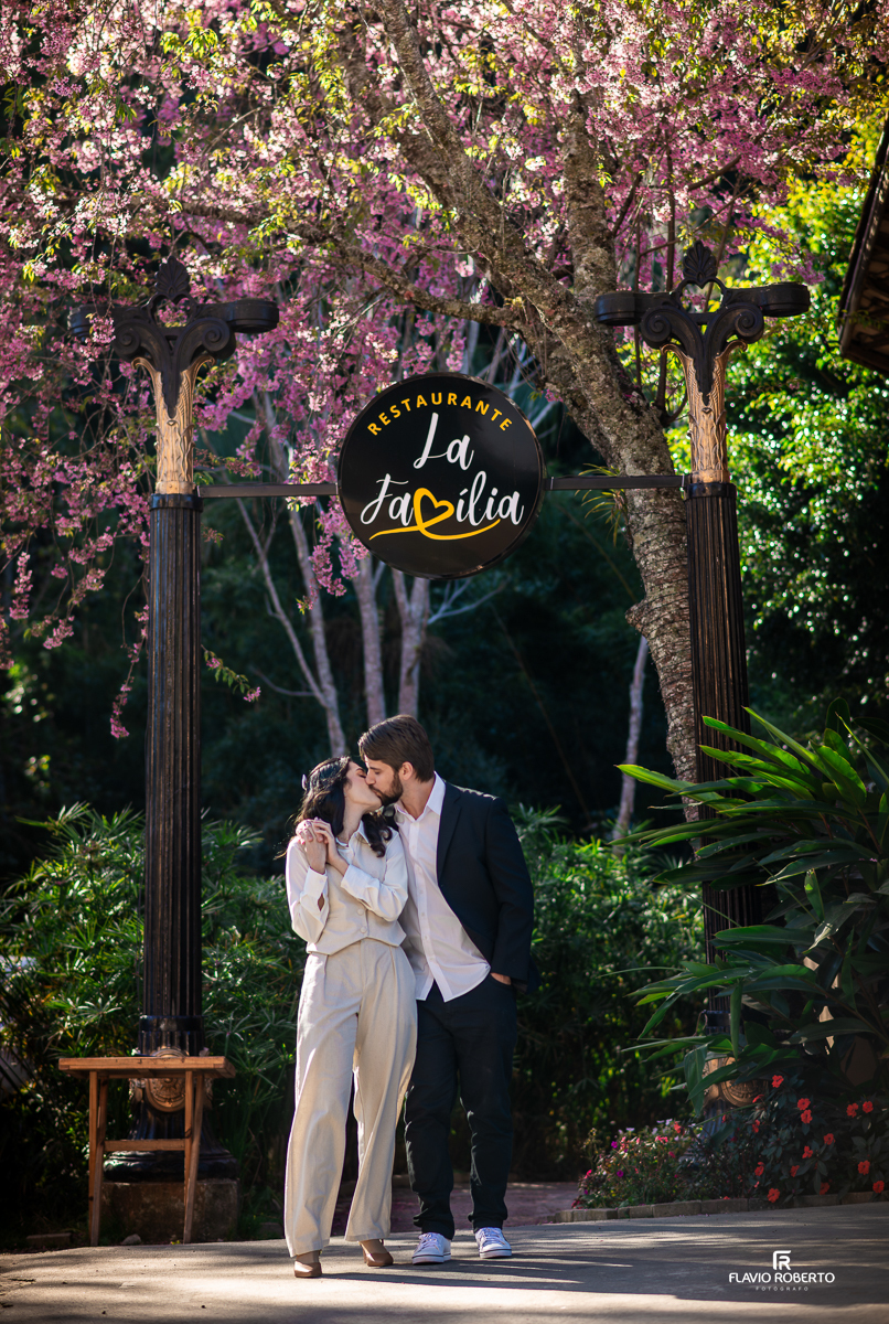 Casal Laís e Gabriel se beijando carinhosamente sob o portal do Restaurante La Família, com cerejeiras floridas ao fundo e clima romântico em Santo Antônio do Pinhal.