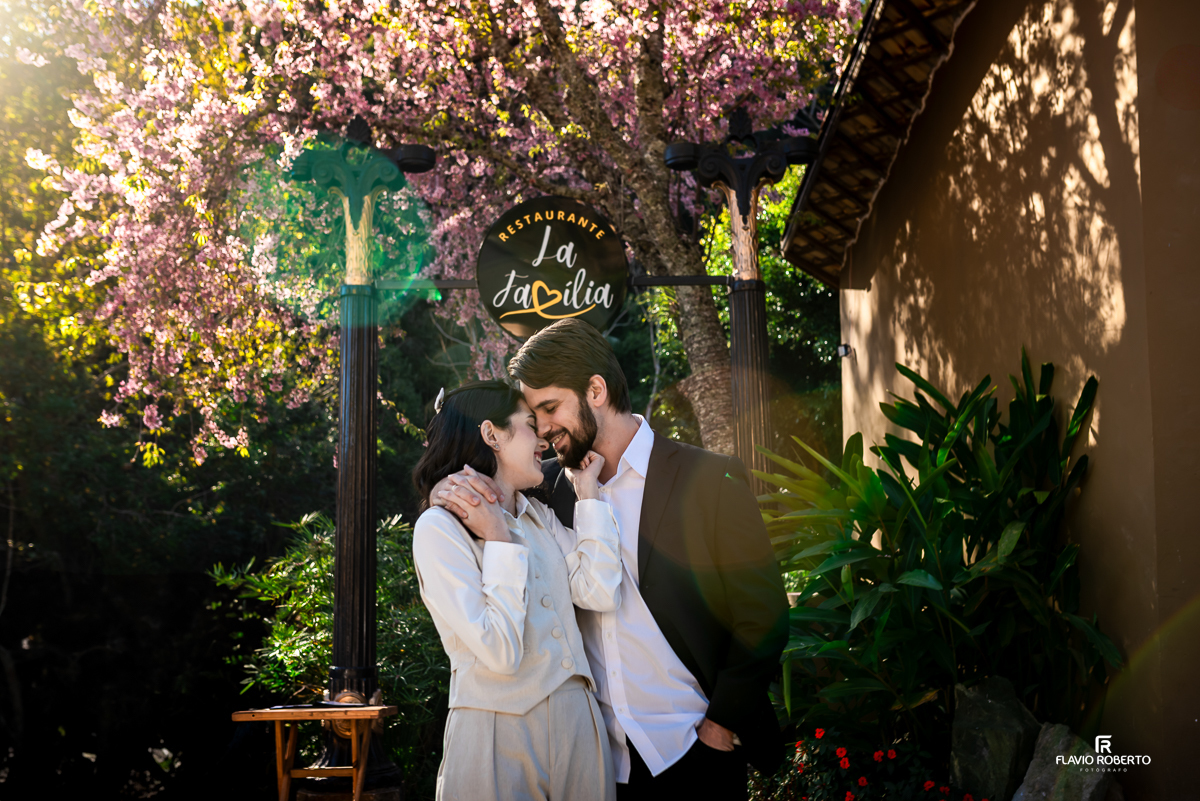Casal Laís e Gabriel se beijando carinhosamente sob o portal do Restaurante La Família, com cerejeiras floridas ao fundo e clima romântico em Santo Antônio do Pinhal.