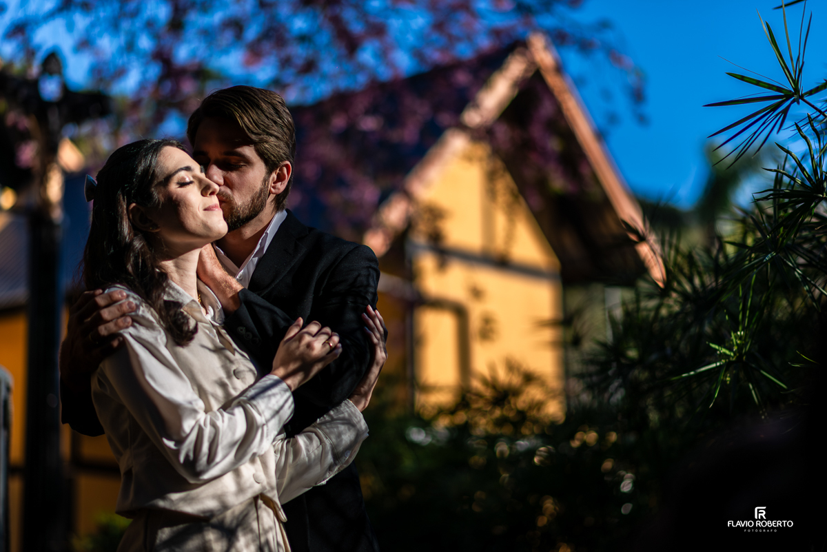 Gabriel beija o rosto de Laís com delicadeza enquanto ela fecha os olhos com expressão de paz e amor, cercados por natureza e arquitetura charmosa.