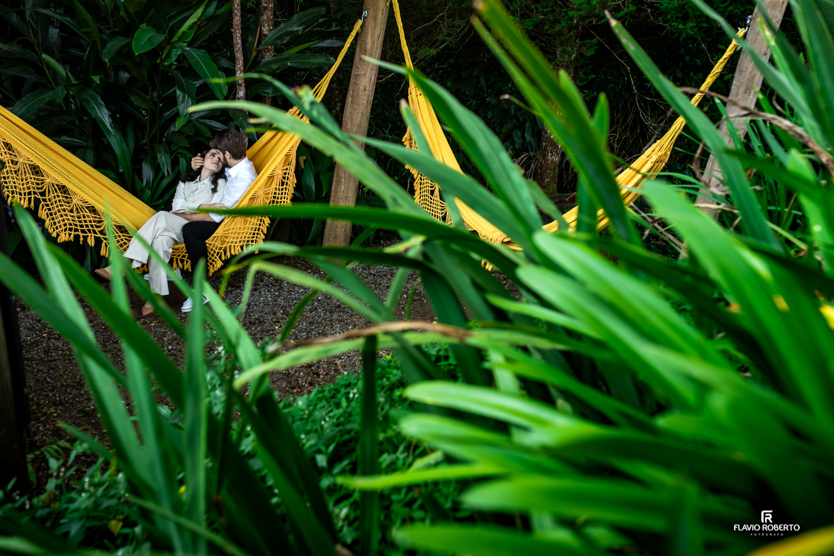 Laís e Gabriel deitados em uma rede amarela cercada por vegetação exuberante, trocando olhares e carinhos em um momento de tranquilidade no meio da natureza.