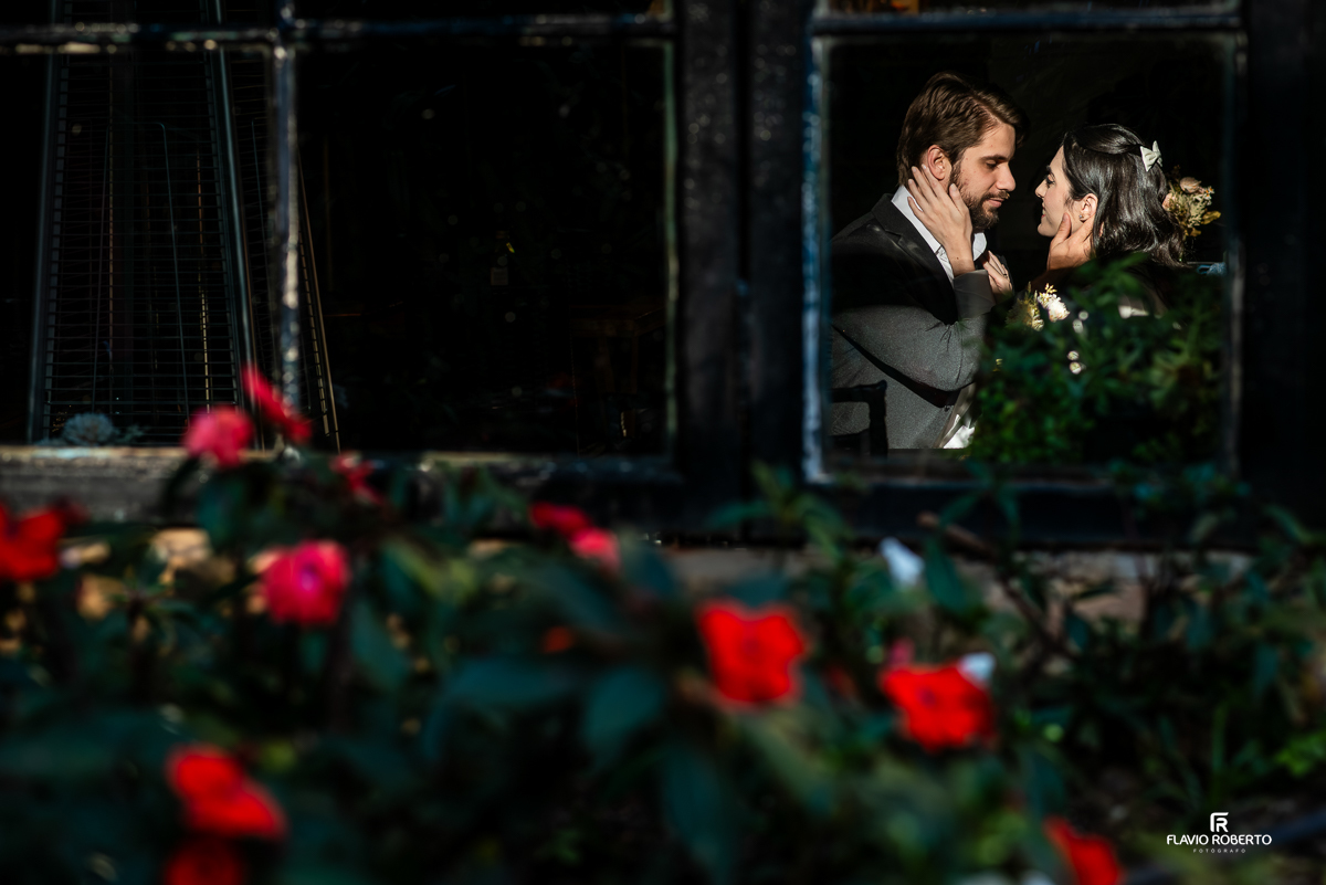 Reflexo romântico de Laís e Gabriel se olhando com carinho, capturado através da janela do restaurante, com flores e lâmpadas vintage compondo a cena.