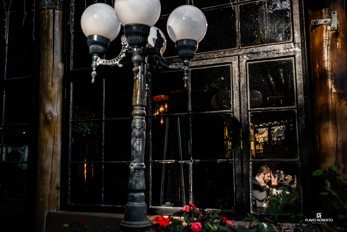 Reflexo romântico de Laís e Gabriel se tocando com carinho, capturado através da janela do restaurante, com flores e lâmpadas vintage compondo a cena.