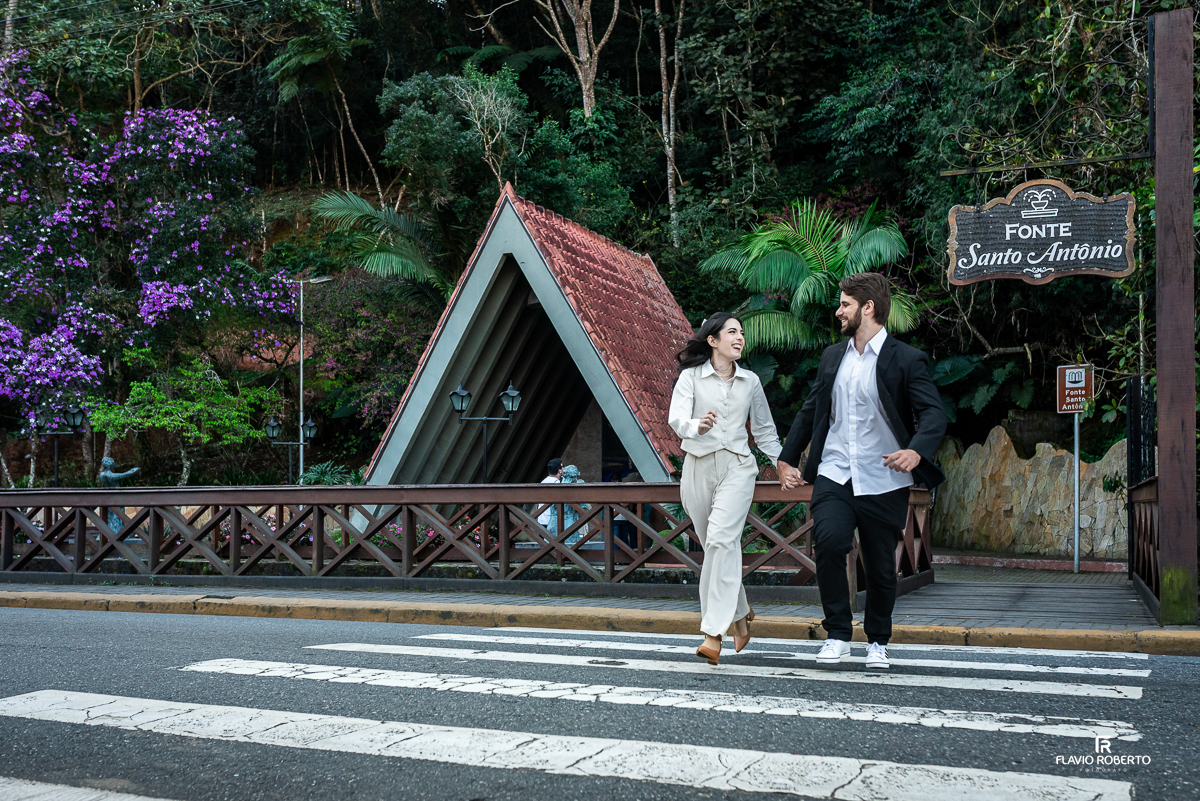 Casal atravessa uma faixa de pedestres de mãos dadas, sorrindo um para o outro em frente à Fonte Santo Antônio.
