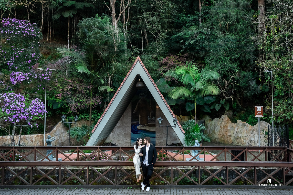 Laís e Gabriel abraçados em frente da Fonte Santo Antônio, rodeados por vegetação exuberante e clima romântico.