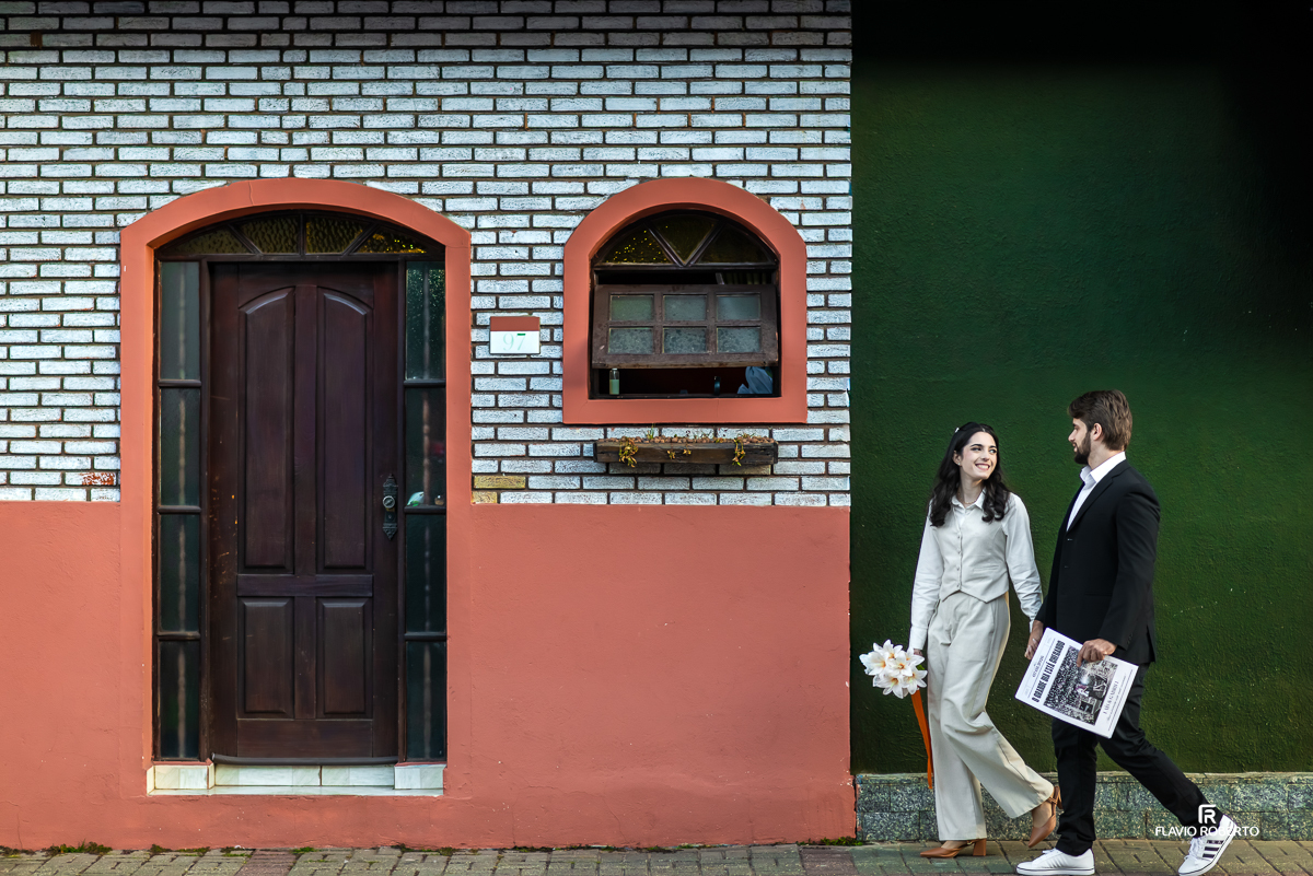 O casal caminha sorridente em frente a uma charmosa casa de tijolinhos brancos com porta de madeira escura no centro de Santo Antônio do Pinhal.